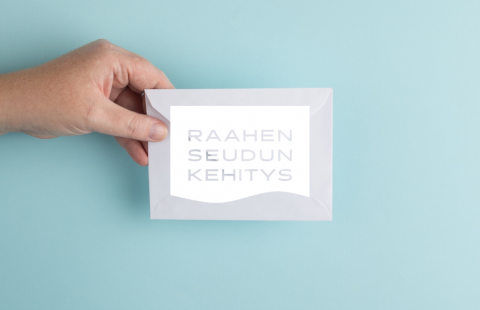 Käsi pitelemässä valkoista kirjekuorta, jossa on Raahen seudun kehityksen logo.